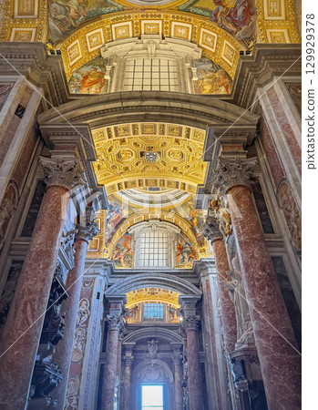 A view looking up inside the majestic St. Peter's Basilica. Massive reddish marble columns frame the ornate ceiling, richly decorated with gilded patterns and detailed religious frescoes 129929378