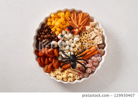 Halloween charcuterie board with pumpkin color chocolate candy, spider, crackers on white background. View from above. 129930407
