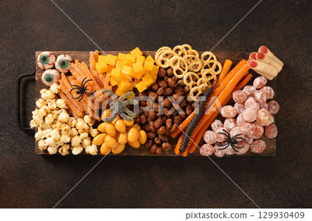 Halloween sweet charcuterie board with pumpkin, popcorn, dried fruits, color chocolate candy, spider, crackers on dark brown background. Festive food for kids party. View from above. Halloween sweet charcuterie board with pumpkin, popcorn, dried fruits, color chocolate candy, spider, crackers on dark brown background. Festive food for kids party. View from above. 129930409