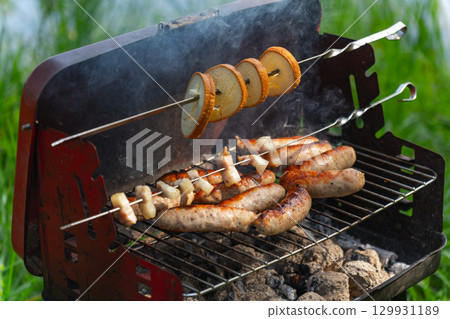 Close-up of a mobile barbecue grill with sizzling sausages, bacon, and bread Close-up of a mobile barbecue grill with sizzling sausages, bacon, and bread 129931189