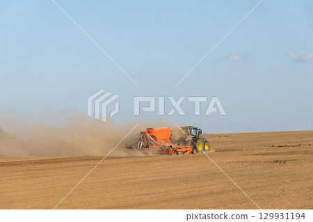 A tractor towing a seeder creates dust clouds while performing field work A tractor towing a seeder creates dust clouds while performing field work 129931194