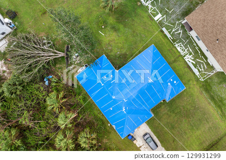 Aerial view of damaged in hurricane house roof covered with blue protective tarp against rain water leaking until replacement of asphalt shingles 129931299