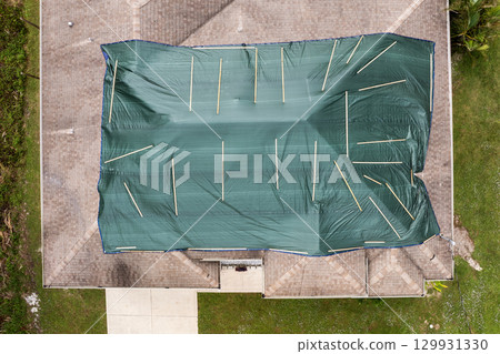Hurricane damaged house rooftop covered with protective plastic tarp against rain water leaking until replacement of asphalt shingles. Aftermath of natural disaster Hurricane damaged house rooftop covered with protective plastic tarp against rain water leaking until replacement of asphalt shingles. Aftermath of natural disaster 129931330