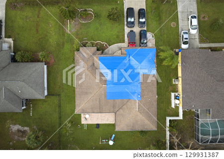 Top view of leaking house roof covered with protective tarp sheets against rain water leaks until replacement of asphalt shingles. Damage of building rooftop as aftermath of hurricane Ian in Florida Top view of leaking house roof covered with protective tarp sheets against rain water leaks until replacement of asphalt shingles. Damage of building rooftop as aftermath of hurricane Ian in Florida 129931387