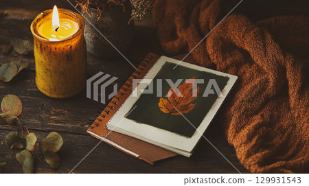 Warm and cozy autumn still life with a lit candle, leaf artwork. A warm, inviting still life with a lit candle, autumn leaf artwork, and cozy knitted blanket. This is AI-generated item 129931543