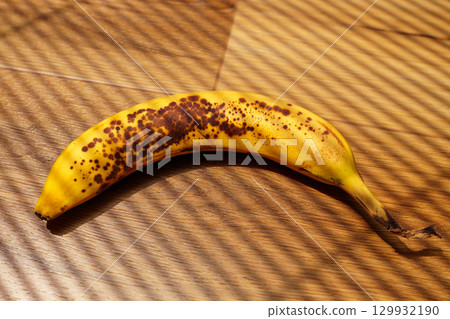 A bright yellow banana with brown spots is placed on a light wooden floor. A bright yellow banana with brown spots is placed on a light wooden floor. 129932190