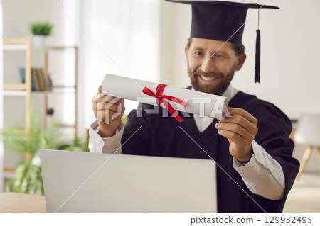 Happy student who has finished online degree is sitting by laptop and holding diploma in hands 129932495