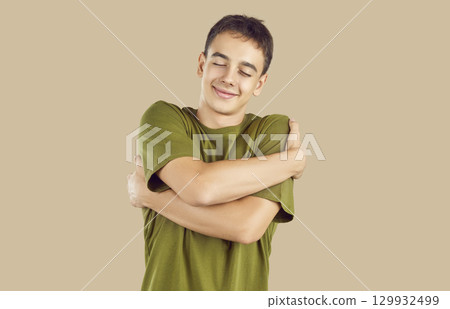 Young teen boy with closed eyes hugging himself isolated on studio beige background. Young teen boy with closed eyes hugging himself isolated on studio beige background. 129932499