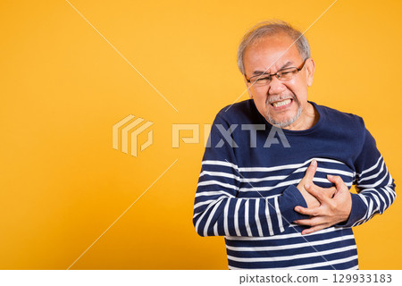 Portrait Asian old man with glasses holds hands on chest studio shot isolated yellow background, senior man pensioner with grey hair heart disease suffering from chest pain 129933183
