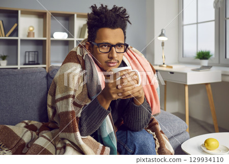 Sad, sick African American man having a cold, sitting on the sofa and drinking hot tea Sad, sick African American man having a cold, sitting on the sofa and drinking hot tea 129934110
