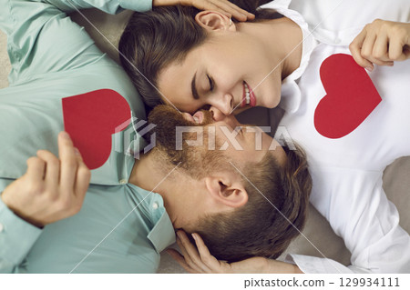 Happy romantic couple lying face to face on floor and holding heart shaped Valentine cards Happy romantic couple lying face to face on floor and holding heart shaped Valentine cards 129934111