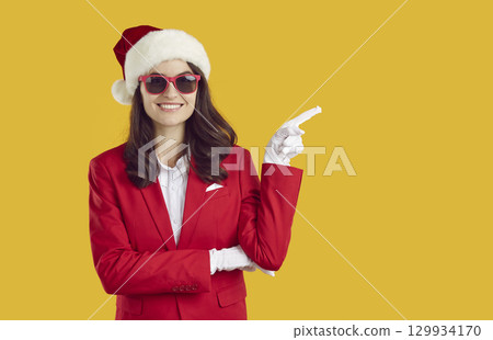 Smiling happy brunette woman in Santa hat and red suit pointing on copy space on yellow background. 129934170