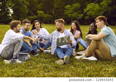 Group of diverse students working together, sitting on grass in university campus and using laptop 129935043