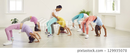 Woman trainer helps flexible children to do bridge exercise at gymnastics studio 129935078