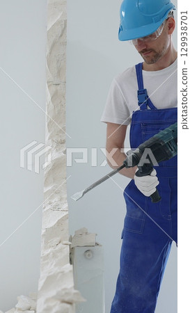 Man construction worker wearing blue overalls, hard hat and protective white gloves, is demolishing white wall with rotary hammer drill, generating dust, close up horizontal view. Renovation concept 129938701