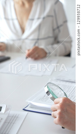 Professional accountants are working together, using a magnifying glass and a calculator, to audit financial reports in office, close up vertical view. Audit and taxes concept 129938712
