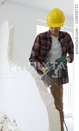 Male construction worker wearing red checkered shirt, yellow hard hat and protective gloves, is demolishing white wall with rotary hammer drill, generating dust, medium long shot. Renovation project 129938715
