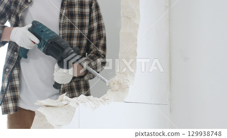 Close up of unrecognizable male construction worker wearing beige checkered shirt and protective gloves, is demolishing white wall with rotary hammer drill, generating dust, close up view. Renovation Close up of unrecognizable male construction worker wearing beige checkered shirt and protective gloves, is demolishing white wall with rotary hammer drill, generating dust, close up view. Renovation 129938748