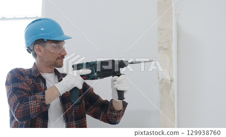 Male construction worker wearing red checkered shirt, blue protective helmet and gloves, is demolishing white wall with rotary hammer drill, generating dust, portrait view. Renovation concept Male construction worker wearing red checkered shirt, blue protective helmet and gloves, is demolishing white wall with rotary hammer drill, generating dust, portrait view. Renovation concept 129938760
