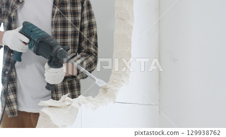 Close up of unrecognizable male construction worker wearing beige checkered shirt and protective gloves, is demolishing white wall with rotary hammer drill, generating dust, close up view. Renovation Close up of unrecognizable male construction worker wearing beige checkered shirt and protective gloves, is demolishing white wall with rotary hammer drill, generating dust, close up view. Renovation 129938762