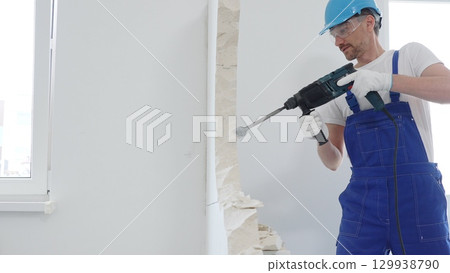Man construction worker wearing blue overalls, hard hat and protective white gloves, is demolishing white wall with rotary hammer drill, generating dust, close up horizontal view. Renovation concept 129938790