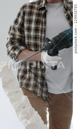 Close up of unknown male construction worker wearing beige checkered shirt and protective gloves, is demolishing white wall with rotary hammer drill, generating dust. Renovation project Close up of unknown male construction worker wearing beige checkered shirt and protective gloves, is demolishing white wall with rotary hammer drill, generating dust. Renovation project 129938793