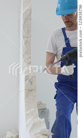 Male construction worker wearing blue overalls, protective helmet and white gloves, is demolishing white wall with rotary hammer drill, generating dust, close up horizontal view Male construction worker wearing blue overalls, protective helmet and white gloves, is demolishing white wall with rotary hammer drill, generating dust, close up horizontal view 129938796