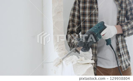 Close up of unrecognizable male construction worker wearing beige checkered shirt and protective gloves, is demolishing white wall with rotary hammer drill, generating dust, close up view. Renovation 129938804