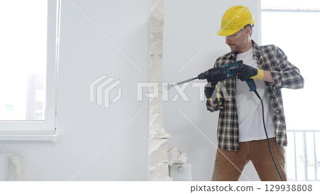 Male construction worker wearing beige checkered shirt, yellow hard hat and protective gloves, is demolishing white wall with rotary hammer drill, generating dust, medium long shot. Renovation project 129938808