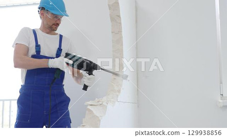 Man construction worker wearing blue overalls, hard hat and protective white gloves, is demolishing white wall with rotary hammer drill, generating dust, close up horizontal view. Renovation concept 129938856