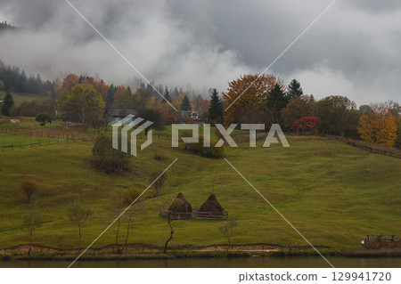 Mountain village near river in mountains on rainy day. Autumn foggy landscape in Carpathian mountains. Tranquil rural landscape with autumn forest, hay stack and local houses. Authentic Ukraine 129941720