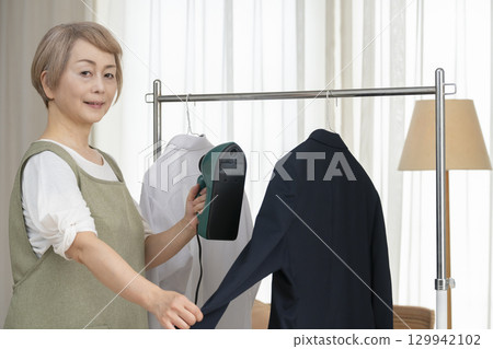 一位老人用蒸氣熨斗熨燙客廳裡掛在衣架上的衣服 129942102