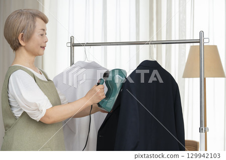 A senior using a steam iron on clothes hung on hangers in the living room 129942103