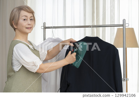 一位老人用蒸氣熨斗熨燙客廳裡掛在衣架上的衣服 一位老人用蒸氣熨斗熨燙客廳裡掛在衣架上的衣服 129942106