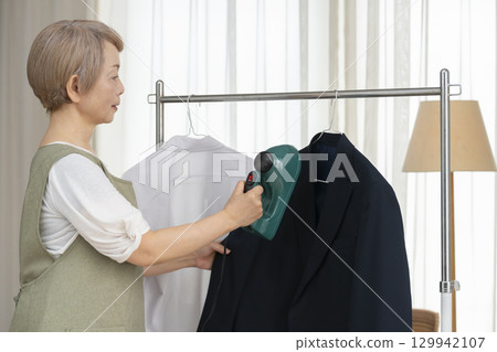 一位老人用蒸氣熨斗熨燙客廳裡掛在衣架上的衣服 一位老人用蒸氣熨斗熨燙客廳裡掛在衣架上的衣服 129942107