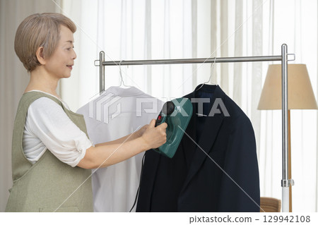 一位老人用蒸氣熨斗熨燙客廳裡掛在衣架上的衣服 一位老人用蒸氣熨斗熨燙客廳裡掛在衣架上的衣服 129942108