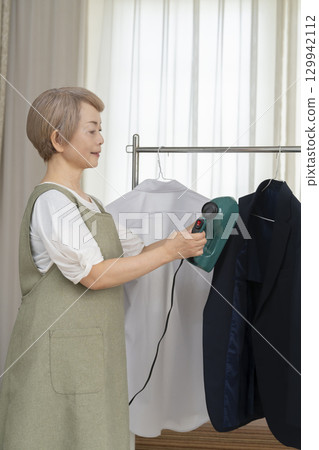 A senior using a steam iron on clothes hung on hangers in the living room 129942112