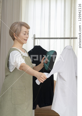A senior using a steam iron on clothes hung on hangers in the living room 129942116