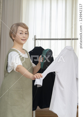 A senior using a steam iron on clothes hung on hangers in the living room A senior using a steam iron on clothes hung on hangers in the living room 129942117