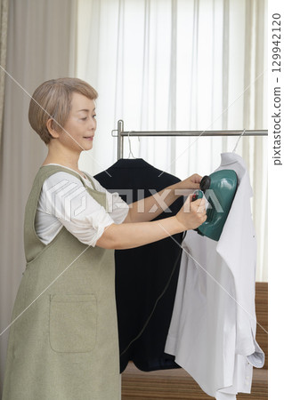 一位老人用蒸氣熨斗熨燙客廳裡掛在衣架上的衣服 129942120