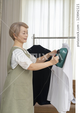 A senior using a steam iron on clothes hung on hangers in the living room 129942121