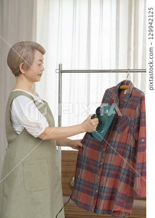 A senior using a steam iron on clothes hung on hangers in the living room A senior using a steam iron on clothes hung on hangers in the living room 129942125