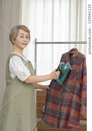 A senior using a steam iron on clothes hung on hangers in the living room 129942126