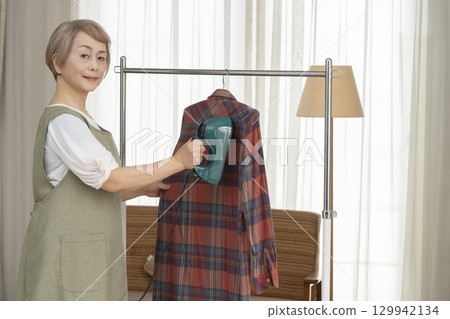 一位老人用蒸氣熨斗熨燙客廳裡掛在衣架上的衣服 129942134