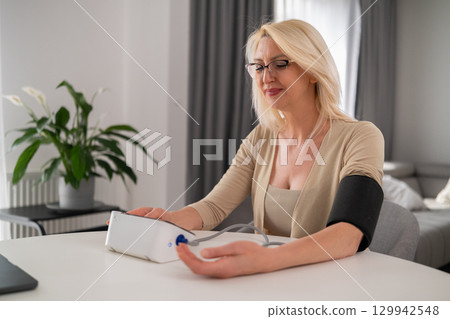 londe woman in beige clothes measuring blood pressure on modern home device, appearing focused and attentive in modern living room londe woman in beige clothes measuring blood pressure on modern home device, appearing focused and attentive in modern living room 129942548
