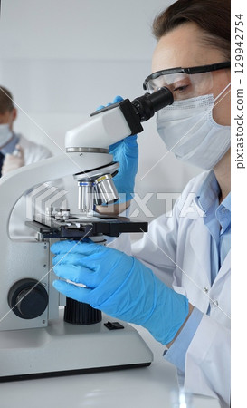 Female scientist wearing mask and blue gloves using microscope doing scientific research in modern laboratory with colleague working in background. Medicine, healthcare and science concept 129942754