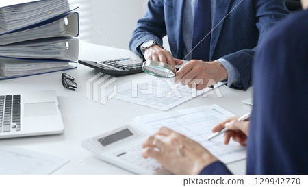 Male financial analysts with female colleague examining documents, using magnifying glass and calculator at workplace in office. Audit and taxes in business Male financial analysts with female colleague examining documents, using magnifying glass and calculator at workplace in office. Audit and taxes in business 129942770