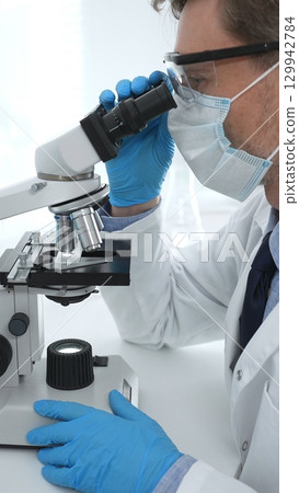 Man scientist wearing protective googles and blue gloves, is using microscope analyzing samples in modern laboratory doing scientific research. Medicine and science concept 129942784
