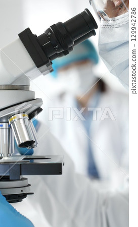 Close up of male researcher examining sample through microscope, wearing blue safety gear in modern laboratory setting. Medicine, healthcare and science concept 129942786