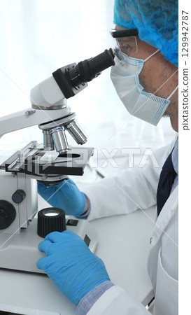 Senior male scientist wearing protective mask, cap and gloves using microscope in laboratory, conducting medical research and analysis. Medicine, health care and science concept 129942787